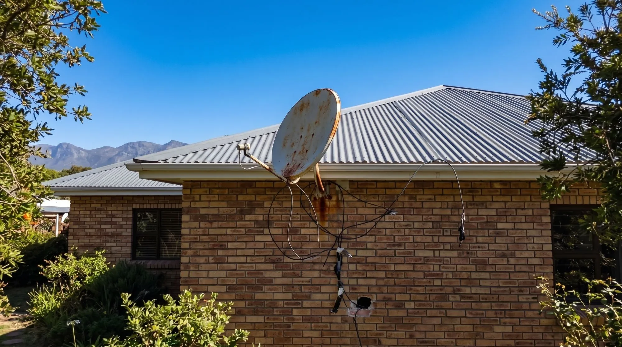 A rusted satellite dish on a face-brick wall below a corrugated roof, with loose cables hanging down the wall